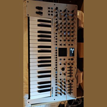 Bílý syntetizátor s klaviatura značky Roland, model GAIA 2. Přístroj má numerous ovládací prvky v podobě posuvníků, otočných knoflíků a tlačítek, vče…